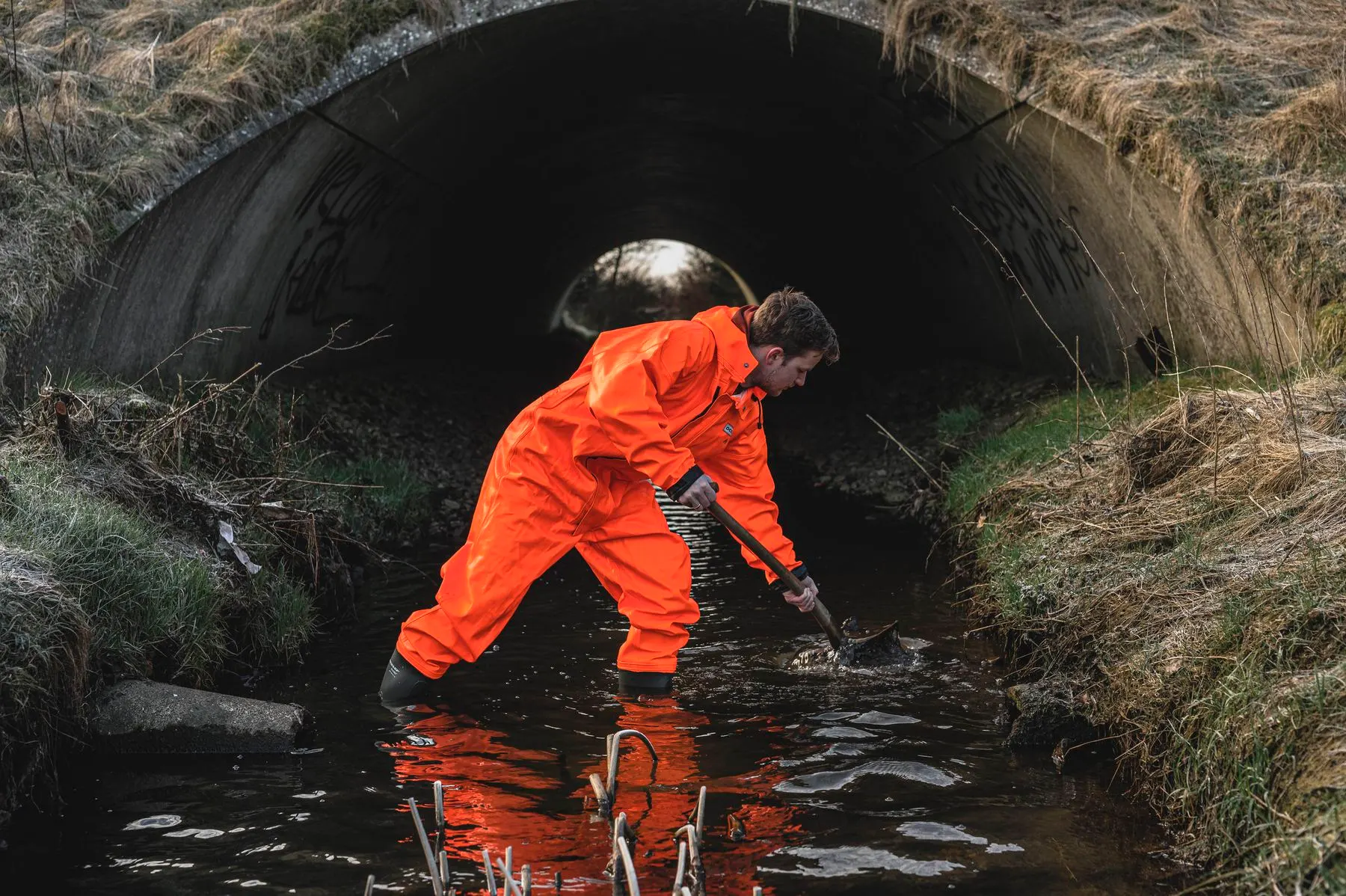 Arbeiter im orangen OCEAN-Regenanzug arbeitet mit Werkzeug in einem Wassergraben vor einem Durchlass